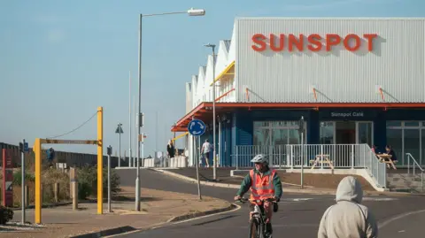 Hufton and Crowe A two-storey building with a white upper floor with pointed roofs and the word SUNSPOT in red lettering on the front. There is a red dividing roof between it and the lower floor, which is dark grey in colour and includes tall windows and doors. A person in a white cycle helmet has just ridden past it.