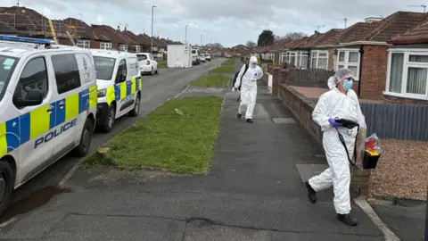 PA Media Several marked police vans are parked along the left side of the road. On the right side of the pavement, two individuals wearing full white forensic suits, gloves, masks and protective shoe coverings are walking in the same direction. One of them is carrying a clear evidence bag and a yellow case. The pavement runs alongside low brick garden walls, and the sky above is overcast with patches of cloud.