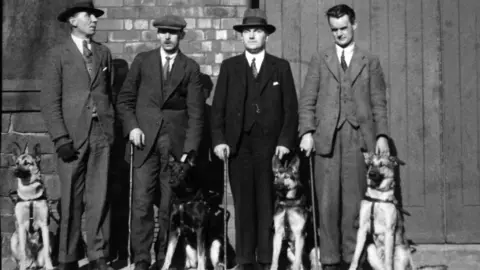 Guide Dogs tbc A black and white image of four men in three-piece suits standing in a line, with four Alsatian dogs by their side. Two of the men are wearing bowler hats, one a flat cap. Three have walking sticks in their hands.