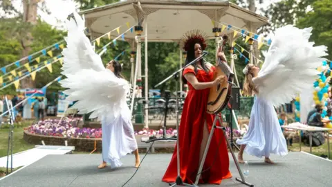 Swindon Welcomes Ukraine A small stage with a bandstand in the background decorated with blue and yellow bunting. There is a woman in a floor-length red dress strumming a lyre-shaped musical instrument and two women wearing white flowing dresses and feathered wings dancing on either side of the musician.