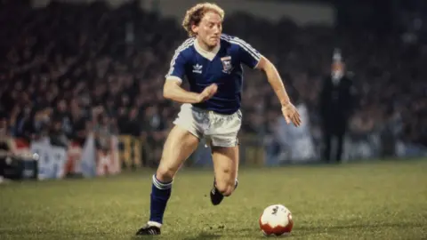 Getty Images Alan Brazil dribbling with the ball at his feet during the UEFA Cup final first leg in May 1981. He is wearing a blue Ipswich Town top with white shorts and blue socks.