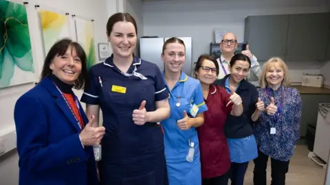 Seven members of staff ( six female and one male) in one of the newly refurbished staff areas. They are all stood smiling at the camera with one of their thumbs up. Four women are wearing traditional hospital scrubs, while two women and one man are wearing traditional office wear. 