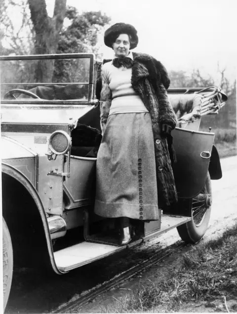 Rolls-Royce A black and white image of Eleanor Velasco Thornton standing on the running board of a Rolls-Royce. She has a jacket over one shoulder and is wearing a jumper and long skirt