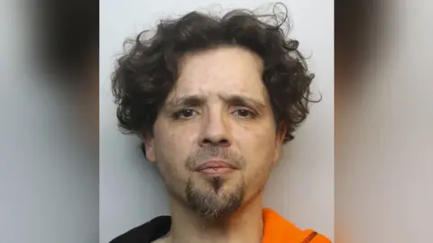 Wiltshire Police A police mugshot of Troy GIbbs, a man in his 40s with dark curly hair, a goatee and lip piercing