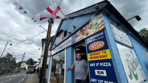 George King/BBC Nuri Ozmicco standing in the doorway of his kebab shop in Framlingham. He is is giving a thumbs up gesture and looking into the camera. Advertisements for food are on the walls of the shop and England flag is flying at the top of the shop. 