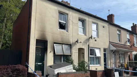 A light-coloured house with some windows out of place. Blue and white police tape is visible on the left and right of the photo.
