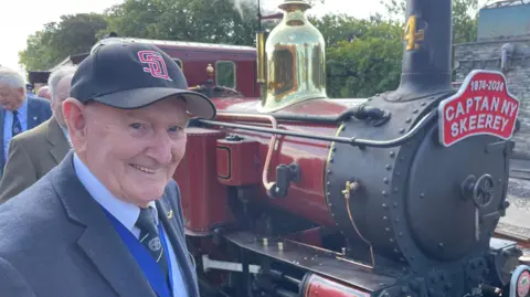 PHIL GAWNE Raymond Gawne, a man with a bright smile, he wears a baseball cap and suit and stands in front of a steam train.