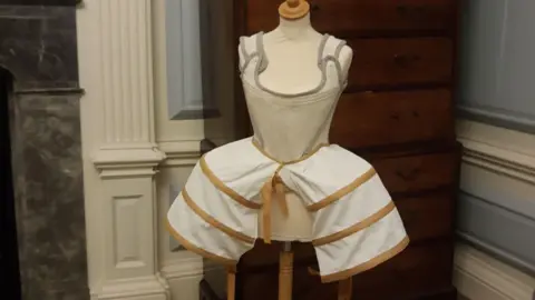 A museum display featuring a historical undergarment on a mannequin. The garment includes a fitted white bodice with grey trim and shoulder straps, attached to a hooped skirt frame with beige bands around it.  Behind the mannequin, there is a wooden chest of drawers and decorative wall mouldings.