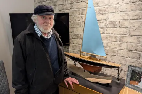 Ian in his living room with a model yacht that is only 3ft tall