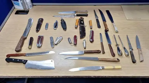 Several knives are on a wooden table. Some of them are small pocket knives, and there are kitchen knives and a meat cleaver.