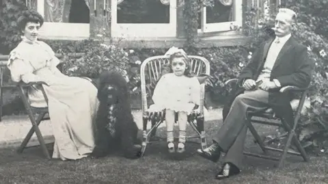 Michelle Bullivant Kate Hall is wearing a long light coloured dress, with her dark hair piled on her head. A large fluffy black dog is sitting at her feet. Next to do the dog is Elsa, aged five to six, sitting on a rattan chair, wearing a short white lace-edged dress with a large light coloured bow on her head. To her right is William Hall, who has short greying hair and a large white moustache. He is wearing a black frock coat over a white shirt, pressed trousers and very shiny black boots. Behind them is a path, with flowers under a bow-shaped window.