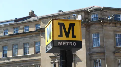 A Tyne and Wear Metro sign. The large yellow cube displays the Metro M logo on a large pole. It is located in Newcastle city centre next to the Monument Metro station.