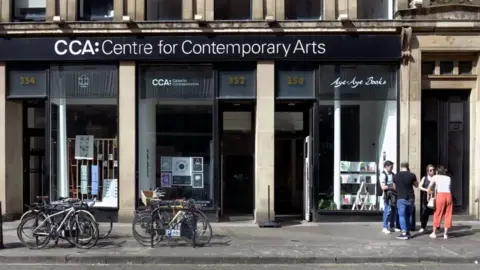 A street view of the Centre for Contemporary Arts building. It has floor-to-ceiling windows and a black banner sign at the top says CCA: Centre for Contemporary Arts. Bikes are locked up outside and a group of people converse on the pavement.
