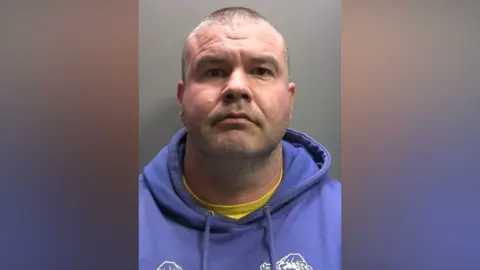 A mugshot of Craig Berry who is staring into the camera with a straight face. He has closely shaved grey and brown hair and is wearing a purple hoodie on top of a bright yellow t-shirt which is just peeking through.