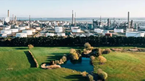 Getty Images An aerial view of the ExxonMobil Fawley oil refinery shows multiple large white round containers and towers overlooking the Solent, with a field in the foreground and a small pond filled with water and surrounded by trees