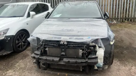 A dark coloured car with a missing bumper covered in plastic sheeting.  