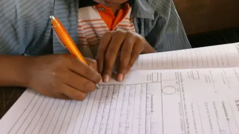 Hands holding an orange pen writing on an exam paper.
