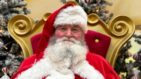 A man dressed as Santa Clause sits on a large gold throne with red velvet cushioning. He has a large white and grey beard, and is smiling at the camera. He also has red cheeks and a pair of small, oval glasses.
