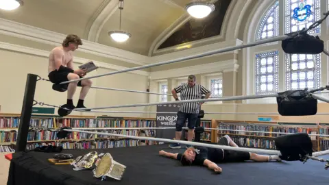 Jessica Bayley/BBC A boy in black shorts and no shirt is sat on the corner of the ropes of a wrestling ring, he is reading a book whilst he sits. There is also a ref stood in the ring as well as a man wearing black shorts and a black T-shirt lying on the floor of the ring. Behind them is a stained-glass window and bookshelves.