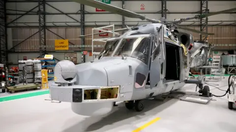 A Wildcat military helicopter with panels missing whilst under maintenance in an aircraft hanger. 