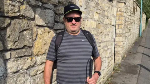 Andy Evans Andy Evans is standing on a pavement in front of a stone wall, wearing a hat and a backpack. In his hand is a white cane, and he is wearing the AI glasses, looking at the camera.