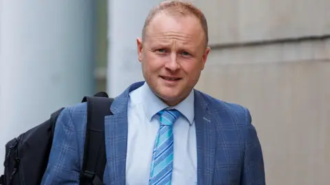 PA A man in a light blue suit with a light blue stripe tie is walking during the day time outside a building. He has thin, balding light hair and is wearing a backpack over one shoulder