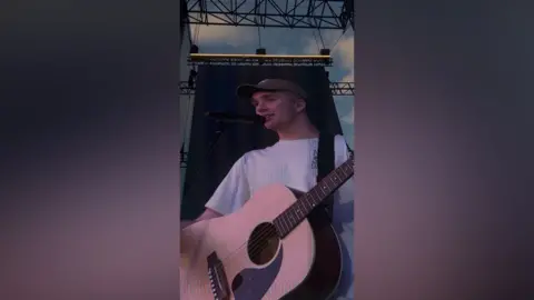 Sarah Kelly The image is a photo taken of a screen at a concert.It shows Paul with a brown acoustic guitar. 