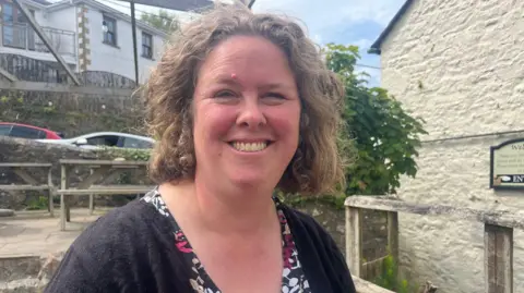 Sarah Treen is smiling as she stands outside the pub. She has shoulder length brown curly hair and is wearing a black cardigan over a black, pink and white printed top. Behind her are parked cars and houses.