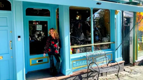 Want That Dress Jane McNally is wearing a black, red and green floral shirt and green trousers. She is standing in the doorway of her shop which is painted light blue with a gold trim.