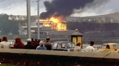 NOEL HOWARTH People on Douglas Promenade standing next to cars from the time looking on as the Summerland complex is engulfed in flames across the bay.