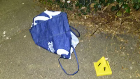 A dark blue reusable Boots carrier bag, with the Boots logo written on it in white lettering, lies on the concrete next to some dried grass and foliage