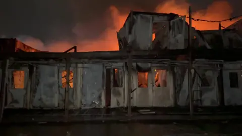 BBC A building on fire at night. It has been badly damaged, with flames and smoke glowing orange behind the shell of the building.