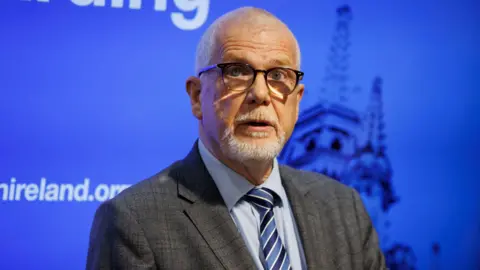 PA Media Convener of the Presbyterian Church in Ireland Rev David Bruce standing in front of a blue TV screen which says Presbyterian Church in Ireland. He has glasses and is wearing a suit