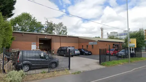 A street view of an industrial estate with vans and cars parked in a gated car park.