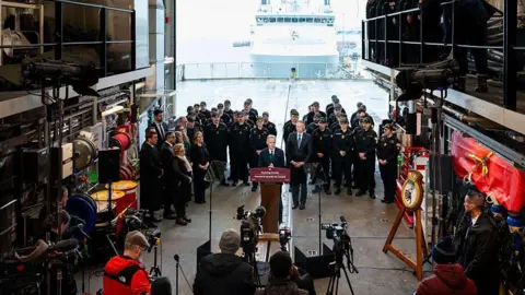 Getty Images A wide shot of a Mark Carney press conference where he is making a defense announcement in Halifax, Nova Scotia. Behind him are members of the Canadian military, and in front of him are TV cameras and journalists. The backdrop is the Halifax shipyard, with a docked large ship visible in the background. 