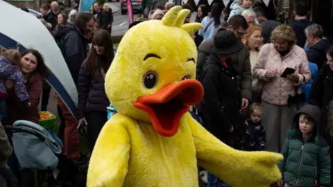 Bradford-on-Avon Town Council Someone wearing a rubber duck suit with a yellow body and orange beak. They are stood with their arms out in a crowd.