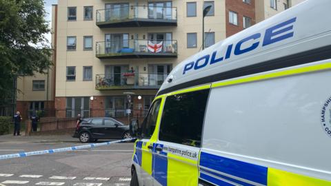 A marked police van parked up on the right side of the picture taken in Chapel Road. Opposite the van is a flat building, where two male police officers dressed in all black uniform stand behind a police tape cordon