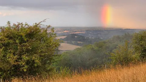 Andy B A small patch of rainbow hovering above a country scene. A field with trees can be seen on the brow of a hill in the foreground with an expanse of countryside and fields that can be seen in the distance.