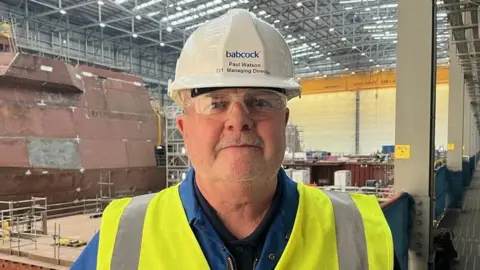 Paul Watson wears a white hard hat with the word babcock on it, and protective glasses. He stands inside a shipyard environment. He is wearing a blue jacket with a neon yellow high vis jacket over it.