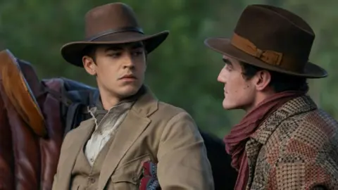 Daniel Smith/Amazon MGM Studios Tiffin wears a brown hat, and beige waistcoat and jacket. Light striped shirt underneath.