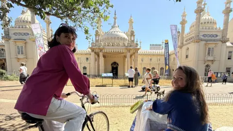 Two women sat in front of Brighton's Royal Pavilion. One is sat at a sewing machine and is working on a piece of art. The other is sat on a bike. Both are looking behind them towards the camera and are smiling.