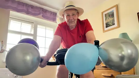 Geoffrey Bolton, 100, on a exercise bike with coloured balloons tied to the handle bars, he is wearing a white fedora hat, black shorts, red top, and glasses. Behind him is a window and pictures on the walls.