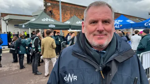 A man with grey hair and grey stubble wearing a GWR coat and fleece underneath is smiling at the camera. He is standing in the car park of a train station where people are queuing.