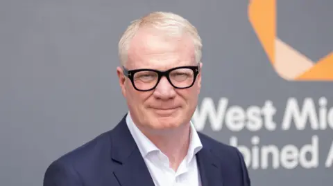 West Midlands Mayor Richard Parker smiles at the camera, in a close-up of his face and shoulders. He stands in front of a grey board which says West Midlands Combined Authority on it.