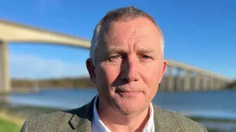 Qays Najm/BBC Mark Ling wearing a green jacket and a white shirt standing in front of the Orwell Bridge