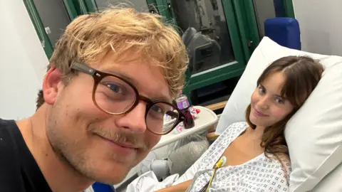 Faye Greenwood A selfie of a blonde man with glasses and a woman with brown hair. The woman is in a hospital gown and is in a hospital bed.
