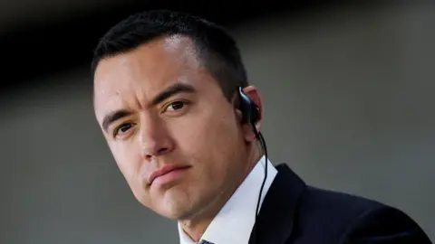 Noboa que há de gravação com um pedaço de ouvido e vestindo um terno escuro, um terno escuro tirado em frente a um fundo cinza Reuters na cabeça tirada em um canto 