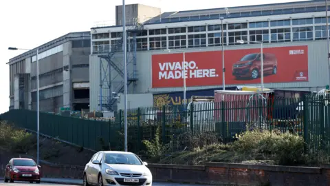 The back of the Vauxhall van plant in Luton, with a huge red poster on the side saying VIVARO MADE HERE and a photo of a red van. Two large industrial buildings stand side by side, with a blue fire escape at the back. Flanked by a tall green metal fence and a road, two cars drive past.