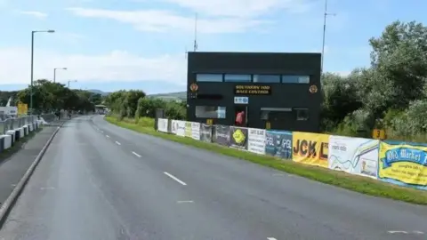 A road with a large black building on the right hand side, barriers are up along the road with different sponsors. 