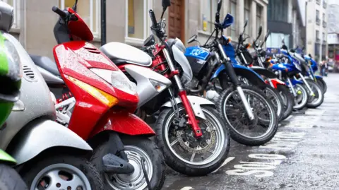 Getty Images A row of motorcycles - some coloured red or blue - parked on a wet street. 